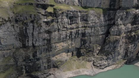 Aerial-pan-down-showing-the-steep-cliffs-towering-next-to-the-Oeschinensee-lake-in-the-Swiss-Alps