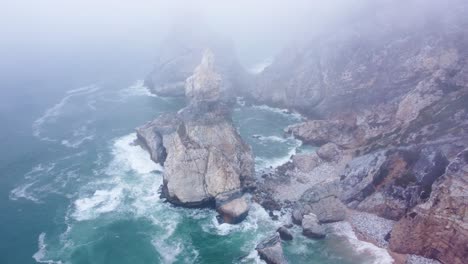 AERIAL-Orbiting-Shot-of-an-Imposing-Rock-in-the-Fog-at-Praia-da-Ursa,-Portugal