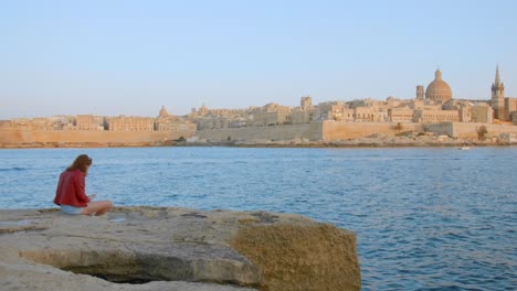 Frau-Liest-Buch-über-Felsiger-Küste-Mit-Blick-Auf-Die-Stadt-Valletta-In-Malta