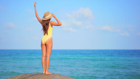 With-her-back-to-the-camera,-a-woman-stands-on-a-boulder-that-overlooks-the-ocean-horizon