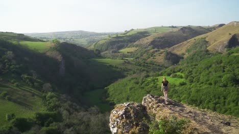 Handheld,-static-wide-shot-of-young-blonde-woman-standing-on-the-edge-of-a-cliff-above-Thor's-Cave,-Peak-District,-England-at-sunset