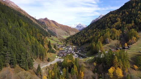 Autumn-view-of-larch-forest-in-Switzerland,-with-a-town,-mountains,-snow-peak-and-a-river-on-the-background