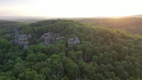 Rocky-cliff-covered-in-lush-forest-trees-with-sunrise-in-horizon,-aerial-view