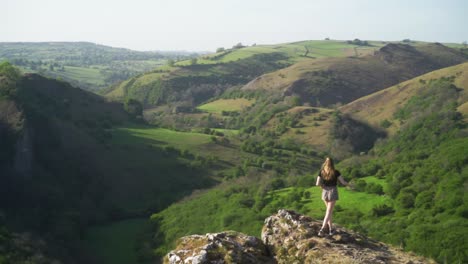 handheld,-wide,-slow-mo-of-young-blonde-woman-standing-on-edge-of-cliff-above-Thor's-cave,-Ashbourne,-Peak-District,-England-at-sunset