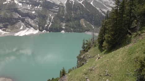 Mountain-reflecting-in-the-water-of-the-Oeschinensee-lake-while-flying-past-a-steep-overgrown-hill-of-fir-and-pine-trees
