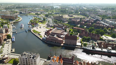 Aerial-view-of-banks-Nova-Motlawa-river-in-Old-Town-of-Gdansk,-tour-ferries-and-big-ship-near-the-quay,-Polish-Baltic-Frederic-Chopin-Philharmonic,-city-Ferris-wheel,-Hotel-KROLEWSKI