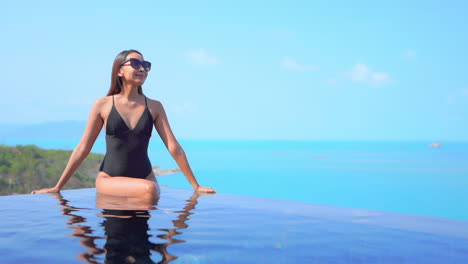 Attractive-woman-in-black-swimwear-and-sunglasses-sitting-on-the-border-of-rooftop-infinity-pool-with-a-turquoise-sea-on-background-leaning-on-the-edge-of-the-pool-on-sunny-day