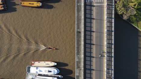 Aerial-birds-eye-shot-of-small-boat-cruising-on-river-under-a-bridge-with-traffic-during-sunny-day