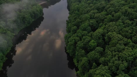 Sky-reflection-in-river-water-surrounded-by-dense-forest,-aerial-view