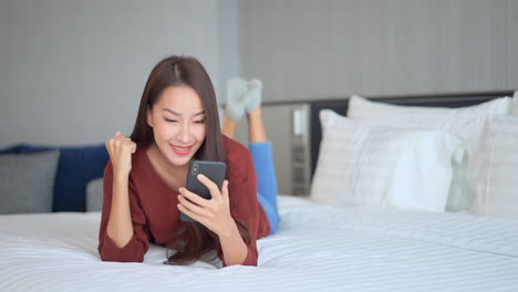 Smiling-young-Asian-woman-lying-on-bed-with-smartphone-in-hand-exults