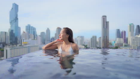 Young-woman-in-white-swimming-suit-inside-rooftop-infinity-pool-on-amazing-blurred-Bangkok-cityscape-background