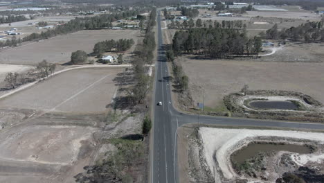 Aerial-wide-shot-that-zooms-in-on-a-white-vehicle-driving-on-a-road-next-to-farms,-in-Stanthorpe-Queensland-Australia