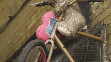 dirty-teddy-bear-left-alone-on-a-bike-mounted-creatively-on-the-wall-with-pink-heart-pillow-saying-mum-emotional-childhood-memories-abandoned-left-alone-grown-up-without-inner-child-slow-motion