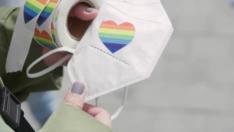 Nahaufnahme-Von-Händen-Mit-Lackierten-Nägeln,-Die-Während-Einer-Pride-Demonstration-An-Einem-Sonnigen-Tag-Einen-Herzförmigen-LGBTQ-Rainbo-Aufkleber-Auf-Einer-Ffp2-Maske-Anbringen