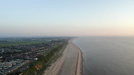 Drone-aerial-shot-flying-along-a-long-sandy-empty-beach-with-grassy-flat-land-on-the-Norfolk-coast-in-Great-Britain-at-sunset