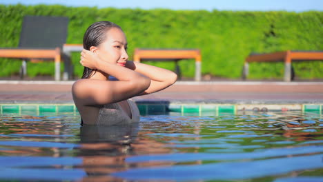 Portrait-of-passionate-young-Asian-woman-touches-her-wet-hair-in-the-swimming-pool-of-the-exotic-hotel-and-turning-her-sight-towards-the-camera