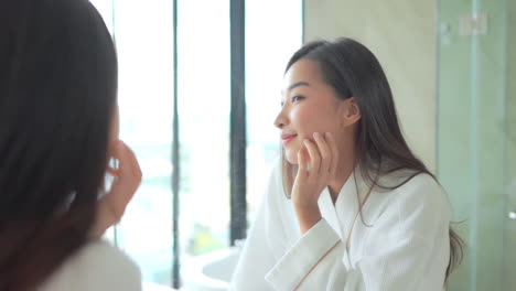 Asian-woman-in-bathrobe-checking-and-touching-her-face-skin-near-the-mirror-smiling-in-the-bathroom-slow-motion-morning