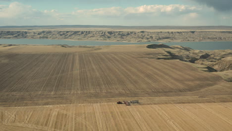 Seeding-Tractor-Working-At-The-Vast-Farmland-With-Calm-Lake-At-Background-In---Aerial-Wide-Shot