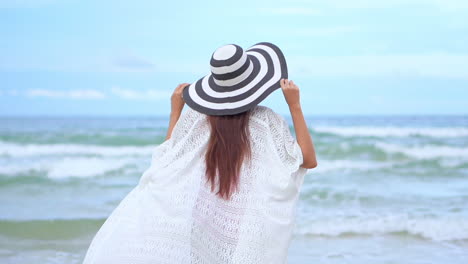 Back-view-of-woman-in-white-dress-and-vintage-hat-on-seashore-in-windy-day