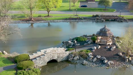 Aerial-of-stone-bridge-to-gazebo-on-island-in-lake,-pond