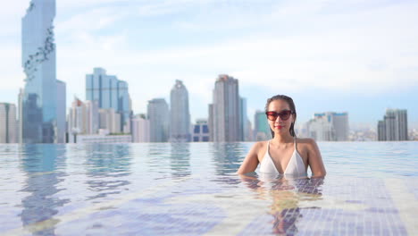A-young-woman,-backed-by-an-urban-skyline,-relaxes-in-a-rooftop-infinity-pool