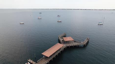 Drone-shot-flying-over-pier-and-blue-water-with-sailboats-in-the-background