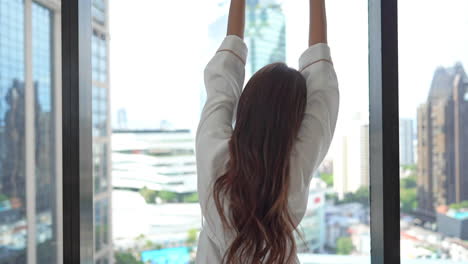 A-young-woman-in-a-bathrobe-and-her-back-to-the-camera-stretches-while-looking-out-the-window-at-an-urban-skyline