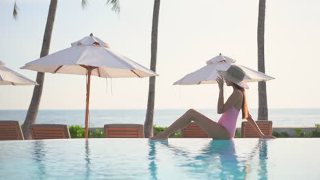 Perched-on-the-edge-of-an-infinity-pool,-a-pretty-young,-fit-woman-in-a-pink-bathing-suit,-sun-hat,-and-sunglasses-leans-back-to-enjoy-and-take-in-her-environment