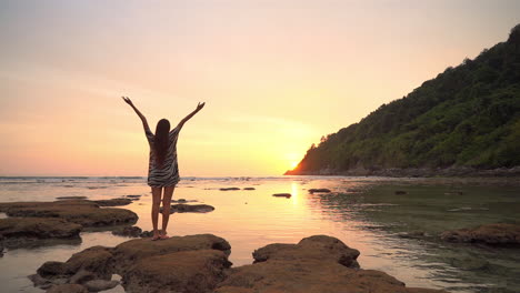 Asian-Woman-standing-on-rock-by-the-sea-water-and-raising-hands-up-when-the-sun-going-down-behind-the-mountain