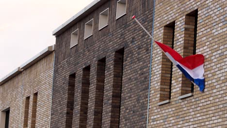Bandera-Nacional-Holandesa-Ondeando-Suavemente-En-El-Viento-Colgando-A-Media-Asta-En-La-Fachada-Exterior-De-Un-Edificio-Moderno-En-Conmemoración-De-Los-Caídos-En-Tiempos-De-Guerra