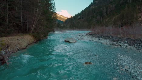 Low-aerial-drone-flight-along-a-scenic-mountain-river-with-fresh-blue-water-in-the-Bavarian-Austrian-alps-in-sunshine,-flowing-down-a-riverbed-along-trees,-forests,-rocks-and-peaks