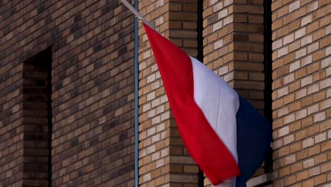 Die-Niederländische-Nationalflagge-Weht-Im-Wind-Und-Hängt-Am-Halben-Mast,-Der-Die-Außenfassade-Eines-Modernen-Gebäudes-Zum-Gedenken-An-Die-Gefallenen-In-Kriegszeiten-Streichelt
