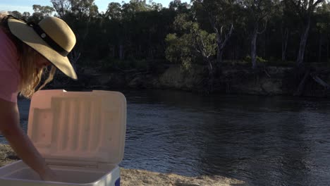 Blonde-woman-checking-what's-in-the-esky-cooler-box-on-river-edge-camping