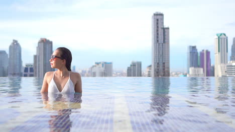 Sexy-Frau-Im-überlaufpool-Auf-Dem-Dach-Auf-Erstaunlich-Verschwommenem-Stadtbildhintergrund-Von-Bangkok,-Handheld-zeitlupe