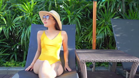 An-attractive-young-woman-in-a-yellow-bathing-suit,-sunglasses,-and-straw-hat-sits-poolside-in-a-sun-lounger