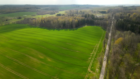 Aerial-orbit-shot-of-hilly-green-farm-fields-surrounded-by-forest-trees-and-driving-road-in-summer