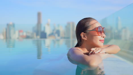 Close-up-of-a-woman-neck-high-in-a-swimming-pool,-rests-her-head-on-her-arms-as-she-looks-out-over-the-modern-city-skyline-Title-space