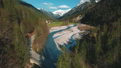 Aerial-flight-over-a-scenic-mountain-river-with-fresh-blue-water-in-the-Bavarian-Austrian-alps-by-afternoon-sunshine,-flowing-down-a-gravel-riverbed-along-trees,-forests-and-hills-seen-by-drone