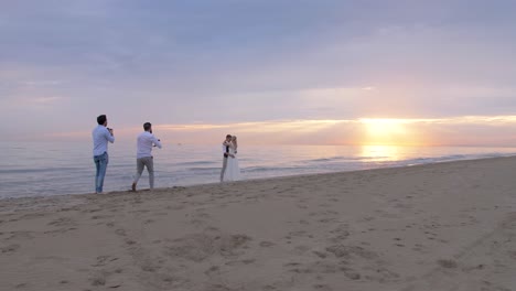 wide-shot-of-the-beach-with-the-sun-setting-over-the-sea,-of-two-photographers-shooting-a-newlywed-couple-during-their-wedding