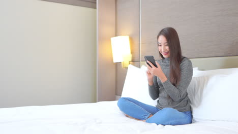Asiatische-Frau,-Die-Mit-Dem-Smartphone-Auf-Dem-Bett-Sitzt-Und-Gute-Nachrichten-Erhält