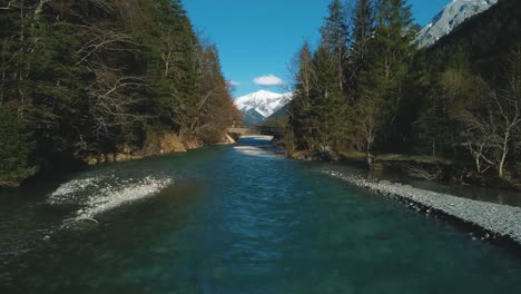 Aerial-flight-along-a-scenic-mountain-river-with-fresh-blue-water-in-the-Bavarian-Austrian-alps-by-afternoon-sunshine,-flowing-down-a-riverbed-along-trees,-a-bridge,-a-street-and-hills-seen-by-drone