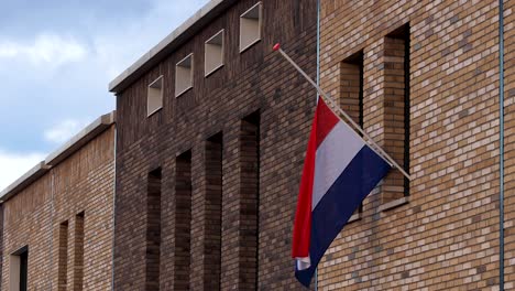 Dutch-national-flag-hanging-half-mast-on-the-exterior-facade-of-a-modern-building-in-commemoration-of-those-fallen-in-war-times-against-an-overcast-blue-sky-in-the-background