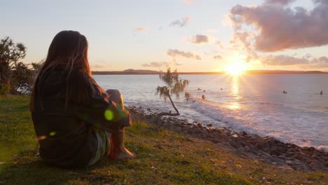 Frau,-Die-Auf-Dem-Gras-Sitzt,-Beobachtet-Die-Surfer-Während-Des-Sonnenuntergangs-Im-Noosa-nationalpark-In-Australien