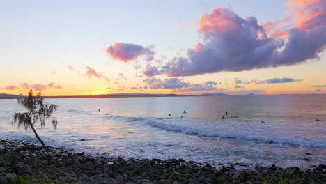 Turistas-Surfeando-En-La-Playa-Ondulada-Con-Costa-Rocosa-Al-Atardecer-En-Queensland