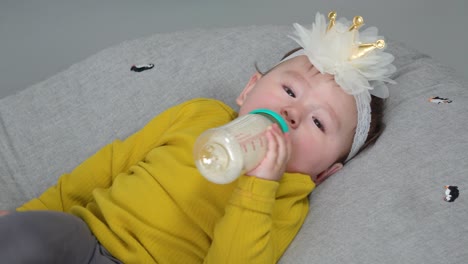 Baby-drink-formula-milk-from-bottle-looking-at-camera-and-holding-the-bottle-in-one-hand-wearing-a-bow-and-yellow-sweater