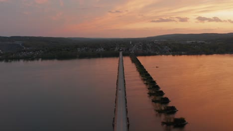 Aerial-of-long-straight-bridge-span-over-wide-river-during-orange-sunrise,-sunset