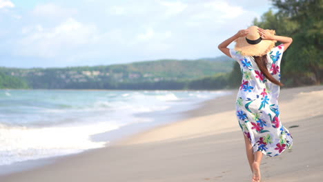Back-view-of-elegant-woman-with-hat-and-floral-dress-walking-on-beach