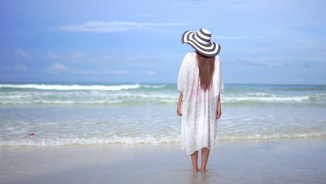 Rear-view-of-barefoot-woman-with-big-hat-and-white-dress-on-beach-looking-sea