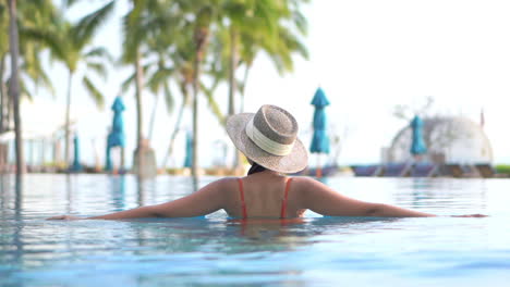 Beautiful-Woman-in-Sunhat-Entering-Swimming-Pool-Water-at-a-Tropical-Island-Resort,-slow-motion-handheld