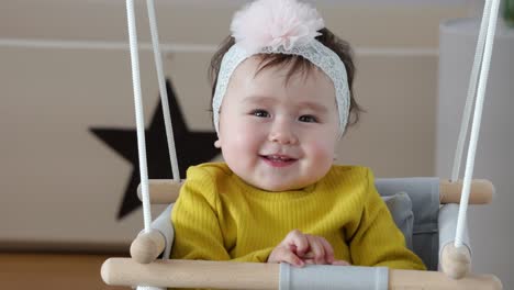 Beautiful-infant-baby-toothy-smile-looking-at-camera-while-hanging-out-in-indoor-swing-at-home-wearing-bow